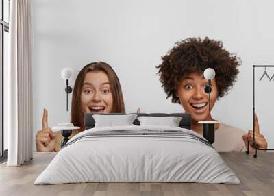 Joyful beautiful mixed race women have satisfied expressions, point upwards, suggest use copy space wisely, dressed casually, pose over white studio wall. Black girl stands near Caucasian friend Wall mural