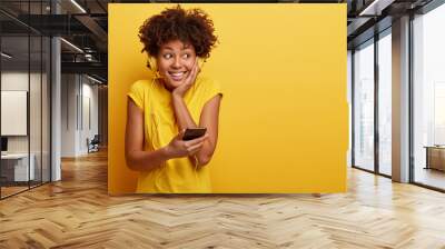 Joyful black woman with toothy smile, listens popular song via headphones, holds modern cell phone, likes listening music during spare time, isolated over yellow background. Teenagers and technology Wall mural