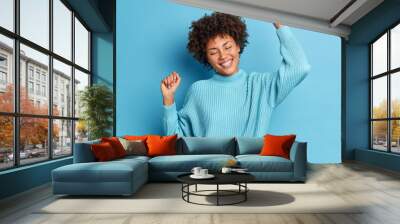 Joyful carefree woman poses full of happiness raises arms and dances with satisfaction smiles pleasantly and closes eyes dressed in long sleeved jumper isolated over blue background has upbeat mood Wall mural