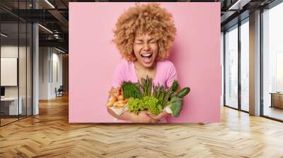 Joyful curly haired woman poses with healthy organic food full of vitamins embraces raw fresh vegetables exclaims from happiness isolated over pink background going to cook vegetarian salad. Wall mural