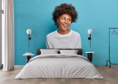 Joyful dark skinned young woman with broad smiles, perfect teeth, feels carefree and enthusiastic, looks happily aside, stands entertained, wears casual white jumper, isolated on blue background Wall mural