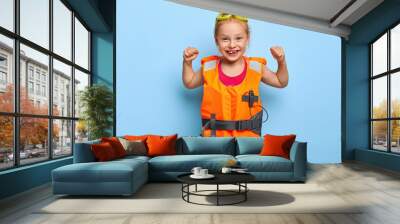 Joyful funny girl with red haired combed in bun, raises arms and shows muscles, wears protective goggles and lifejacket, ready for swimming in pool or sea, isolated on blue background. Children, rest Wall mural