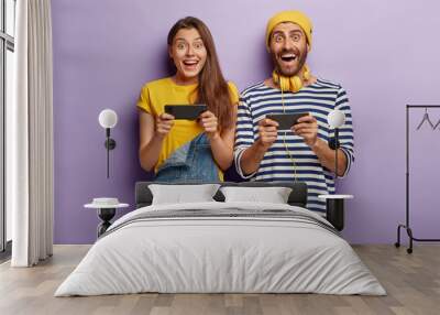 Joyful happy woman and man play games on smartphone, challenge each other, browse net, use headphones for listening playlist, stand closely to each other. Relationship and technology concept Wall mural