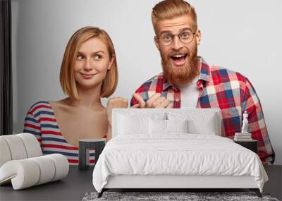 Joyful happy young couple have fun together, indicate at each other, have happy expressions, stand against white background. Glad European woman with bobbed hairdo and bearded ginger guy indoor Wall mural