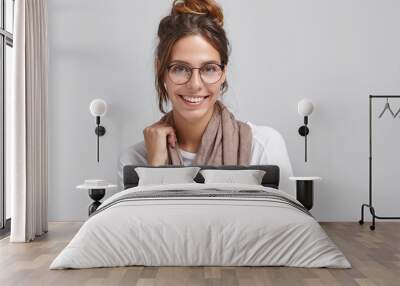 Joyful successful young European female artist or worker of creative profession wearing stylish round glasses and scarf around her neck, looking at camera and grinning broadly, showing white teeth Wall mural