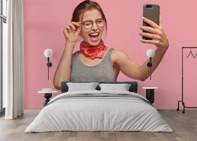 Joyful young lady blinks eyes, makes photo of herself with mobile phone, video call, glad to communicate with friend, wears big round spectacles, poses at studio. Its time for selfie. Leisure Wall mural