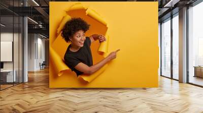 Joyous friendly looking smiling girl points aside with happy expression, toothy smile, pleased to show awesome advertisement, dressed casually says use copy space wisely gestures through ripped paper Wall mural