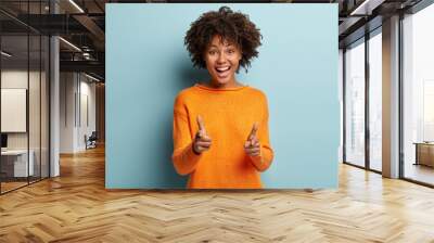 Joyous pretty African American lady makes finger gun gesture at camera, expresses choice, smiles broadly, dressed in orange jumper, isolated over blue background, says you are chosen. Bang, bang Wall mural