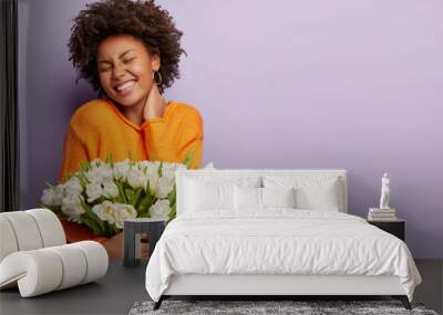 Jubilant upbeat black woman has curly hair, broad shining smile, keeps hand on neck, eyes shut, wears orange sweater, holds white flowers with pleasant scent, models over purple studio wall. Spring Wall mural