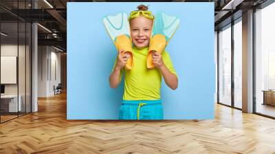 Little girl swimmer wears goggles, holds flippers, has broad smile, lack of teeth, wears green t shirt and shorts, enjoys summer rest, active games on water, isolated on blue wall. Happy childhood Wall mural