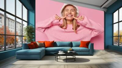 Look at my shining smile. Positive fair haired European woman points at her white teeth smiles broadly wears fashionable panama and casual jumper isolated over pink background has happy mood Wall mural