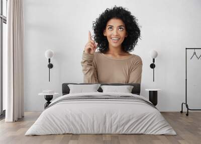 Look at that! Indoor studio portrait of cheerful attractive casually dressed young female with curly hair pointing her index finger up, indication something interesting, having happy excited look Wall mural