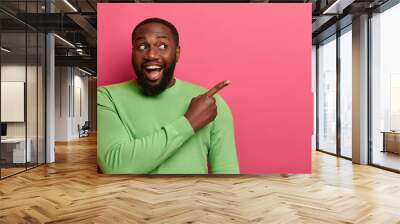 Look attention! Black bearded man points index finger aside, shows copy space on right, empty place for your promotional text, wears green jumper, isolated over pink background. Studio shot. Wall mural