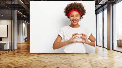 Love to you, my dear. Glad satisfied African American woman makes heart gesture, smiles pleasantly at camera, wears casual outfit, expresses good feelings to lover, poses indoor over white background Wall mural