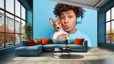 Lovely African American woman carries small breed dog near face keeps lips folded as if wanting to kiss someone loves her favorite pet have walk dressed casually listens music in stereo headphones Wall mural