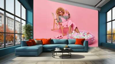 Lovely curly haired woman takes selfie makes peace gesture shares photos with friends in social networks enjoys summer time poses on deck chair keeps legs in inflated pool isolated on pink background Wall mural