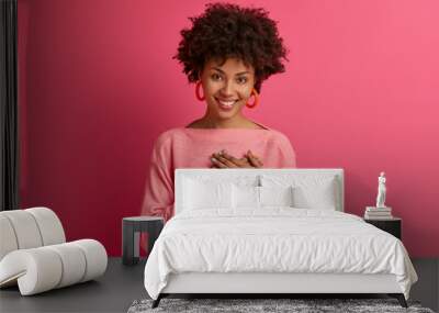 Lovely curly young woman presses palms to heart, feels touched and grateful, looks with admiration at something, keeps all good memories, dressed in pink jumper, isolated over rosy background Wall mural