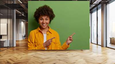 Lovely dark skinned woman with Afro hair points fore fingers right, suggests going this direction, demonstrates awesome product, wears yellow jacket, isolated on green background, has positive smile Wall mural