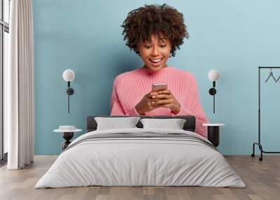 Lovely happy African American woman holds modern cell phone, glad to get message from boyfriend, types feedback, checks balance online, wears pink oversized sweater, stands over blue studio wall Wall mural