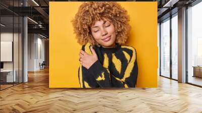 Lovely happy curly haired woman embraces herself daydreamsand embraces herself with both hands recalls romantic event wears comfortable jumper isolated over yellow background. Egoism concept Wall mural