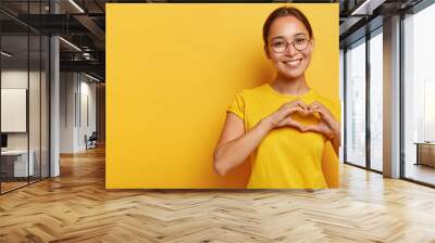 Lovely korean woman with happy smile, shapes heart with both hands, expresses love to you, wears round spectacles and yellow t shirt, says be my valentine flirts with boyfriend poses indoor copy space Wall mural