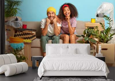 Lovely spouses of different race sit closely on comfortable sofa, being happy apartment owners, have to bring house in order after relocation, unpack boxes, rejoice successful expensive purchase Wall mural