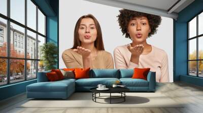 Lovely young Caucasian and African American women blow air kiss at camera, express love to other people, say goodbye on distance, dressed in casual outfit, pose together against white background Wall mural