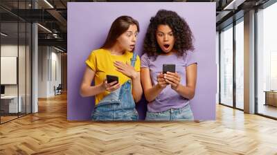 Millennial generation people look with shocked expressions at cell phone, network online, read message with bad surprising content, wears stylish clothes, pose closely over purple background Wall mural