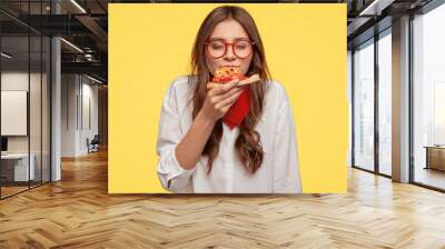 Mmm, so delicious! Dark haired pretty woman eats slice of Italian pizza, keeps eyes closed from pleasure, enjoys nice taste, wears glasses and shirt, isolated over yellow background. Eating concept Wall mural