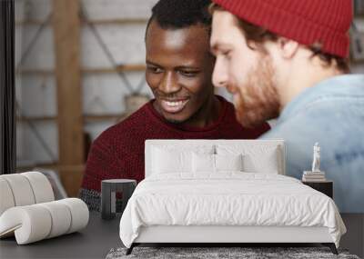 Modern technology, people, leisure and online communication. Happy young dark-skinned male holding cell phone and showing something to his stylish Caucasian friend, both looking at screen and smiling Wall mural