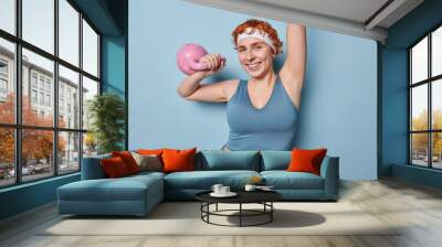 Motivated athletic redhead woman lifts weight works out in gym or at home being full of energy gets fit dressed in tracksuit has workout with coach motivates you to do sport poses over blue background Wall mural