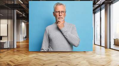 Old age and thoughts concept. Serious grey haired man holds chin and concentrated with pensive expression somewhere dressed in casual turtleneck isolated over blue background thinks over problems Wall mural