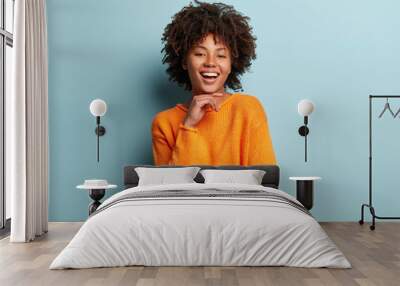 Optimistic Afro American woman with dark skin, curly hair, keeps hands partly crossed, looks at camera with pleasure, wears orange sweater, isolated over blue background. Happy emotions concept. Wall mural