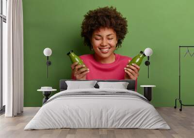 Optimistic dark skinned woman with curly hair holds two bottles of spinach smoothie, has healthy proper nutrition, dressed in casual pink jumper, isolated on green background. People, dieting concept Wall mural