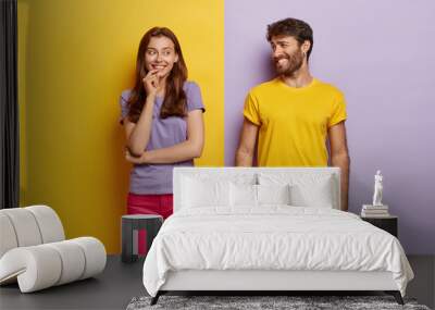 Optimistic young female and male with broad smiles, feel happy, dressed in casual clothes, have fun together, stand against purple and yellow background. People, relations, togetherness concept Wall mural