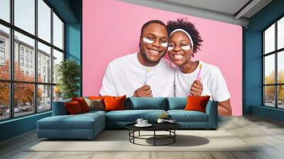 Oral hygiene concept. Positive dark skinned Afro American woman and man hold toothbrushes apply beauty patches under eyes dressed in casual white basic t shirts isolated over pink background. Wall mural