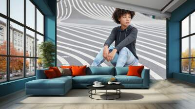 Outdoor shot of pensive dark skinned teenager dressed in casual sportsclothes, sneakers, sits on asphalt, recreats after having sport competitions, has fit body. Teenagers and active lifestyle concept Wall mural