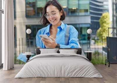 Outdoor shot of positive Asian woman holds cellular gadget and takeaway coffee checks received notification wears spectacles and blue shirt poses against urban background creats social publication Wall mural