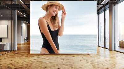 Outdoor view of pleasant looking young female model in bikini and straw hat looks with dreamy expression away, enjoys beautiful ocean view and sunrise, spends summer holiday at beach in tropics Wall mural