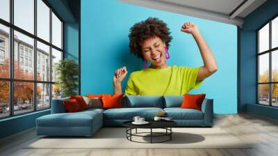 Overjoyed African American woman makes winning gesture expresses happiness raises arms and dances carefree celebrates victory dressed in t shirt isolated over blue background. Female triumphant Wall mural