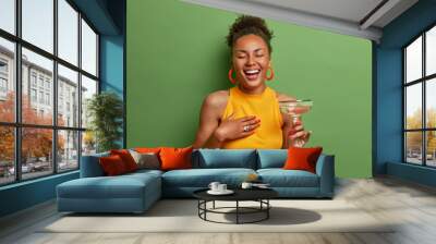 Overjoyed dark skinned woman laughs happily keeps eyes closed and smiles broadly has fun at party drinks cocktail expresses positive emotions wears yellow shirt poses over vivid green background Wall mural