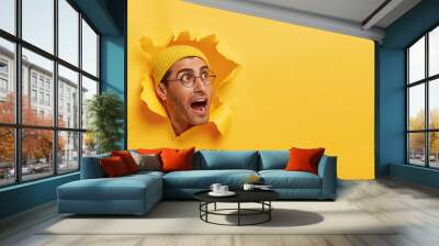 Overwhelmed young guy keeps head through torn hole in yellow background, keeps jaw dropped, gazes aside on free space, notices something surprising, wears hat and big round glasses. Reaction Wall mural