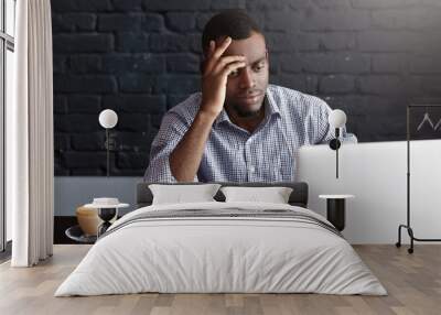 Overworked concentrated young African-American copy-writer wearing shirt working on new articles using laptop computer, leaning his elbow on cafe table with notebook pc, papers, mug and phone Wall mural