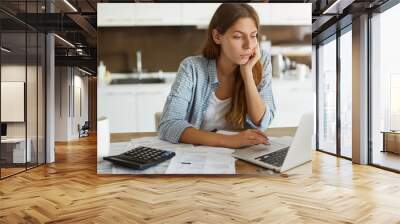 People, accounting, finances, family budget and financial issues concept. Serious young European woman calculating domestic expenses, sitting at dining table in front of open laptop computer Wall mural