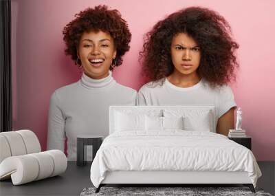 People, emotions and friendship concept. Lovely cheerful Afro woman smiles happily, has carefree face expression wears white turtleneck stands near dissatisfied gloomy female friend upset by something Wall mural