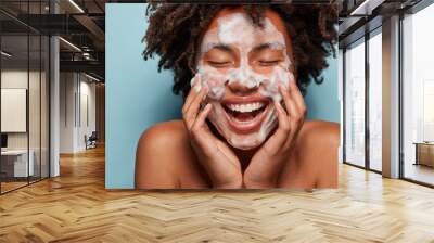 People, skin care and rejuvenation concept. Pleased dark skinned woman with soft skin, applies white foam on face, touches cheeks, smiles broadly, cares of hygiene, feels relaxed and satisfied Wall mural