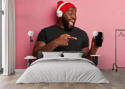 People, technology, lifestyle and advertising concept. Happy dark skinned man shows blank screen of smartphone device, listens audio track in wireless headphones, isolated on pink background. Wall mural