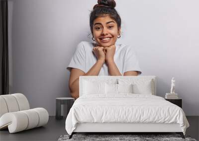 People and happiness. Indoor photo of young fit beautiful cheerful smiling broadly European girl wearing casual clothes posing isolated in centre on white background keeping raised hands under chin Wall mural