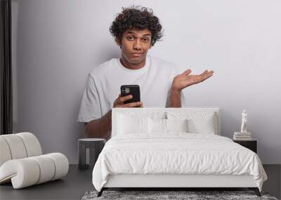 People emotions concept. Indoor photo of young confused Hindu man standing isolated in centre on white background holding smartphone can't find necessary information on internet wearing t shirt Wall mural