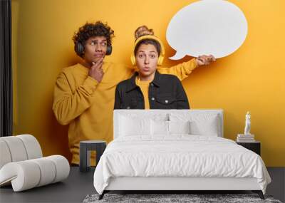 People emotions concept. Studio shot of young thoughtful Hindu man looking and holding white speech bubble with blank space for promotion and surprised European girl standing close to each other Wall mural